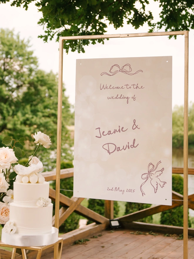 a gold frame with a hanging sign in the midlle which has wording welocme to the wedding of Jeanie & David and the wedding date and on the top is whimsical bow and in the corner is a dove carrying a bow, at the side of the stand is a white wedding cake with a boquet of white flowers
