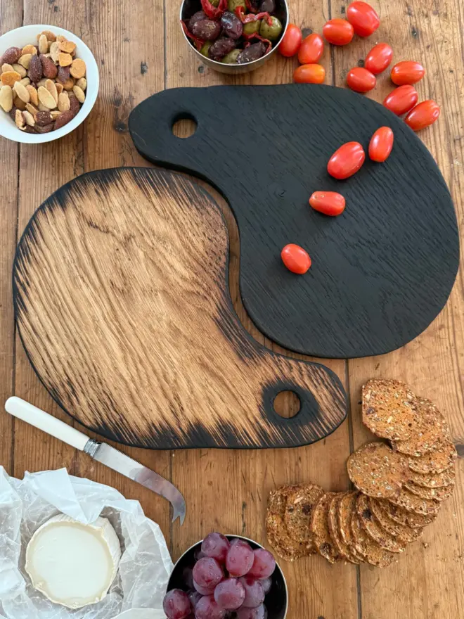 “Pair of handmade Yaki Sugi serving boards in blackened and natural oak, styled together with olives, cherry tomatoes, grapes, crackers and cheese on a rustic wooden table.”