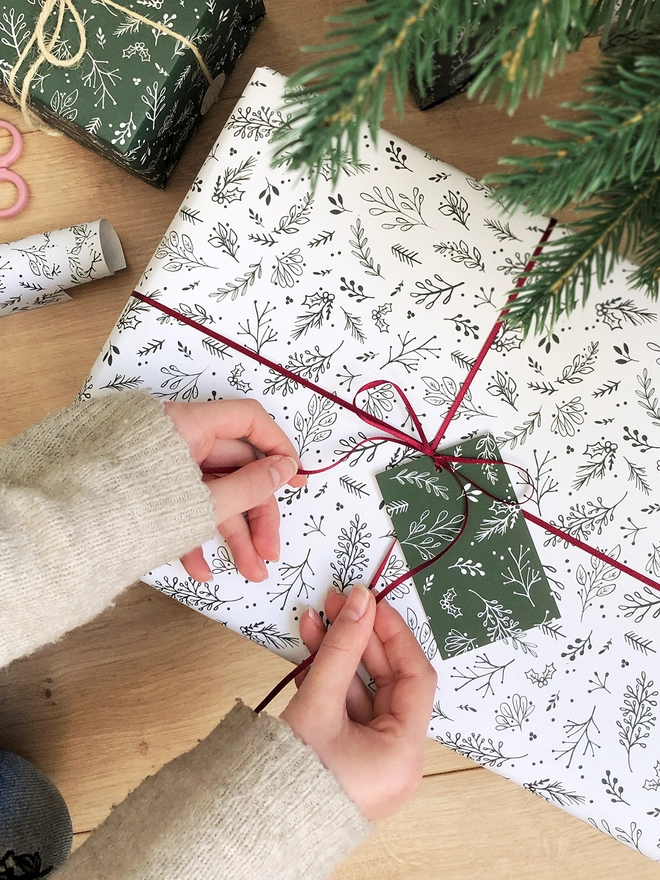 A pile of gifts wrapped in white wrapping paper with a green foliage design lay on a wooden floor beneath a Christmas tree, while hands are tying a red ribbon around one.