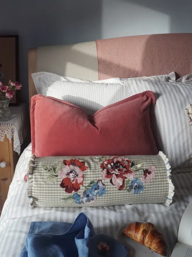 Embroidered floral bolster cushion in sage green gingham, styled on a bed with pink velvet and striped cushions.