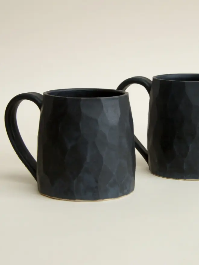 faceted matte black ceramic mug. pair of mugs photographed against off-white studio background.