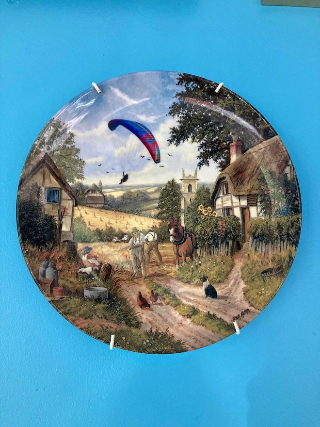 China plate featuring a rural farming scene with the addition of a modern paraglider. 