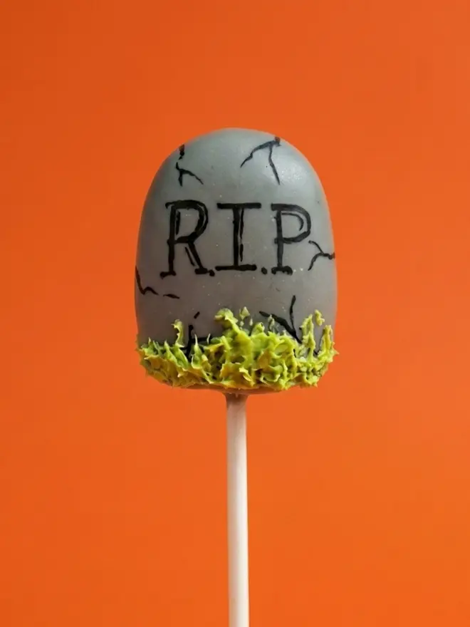 RIP grey gravestone cake pop on a white stick against a bright orange background