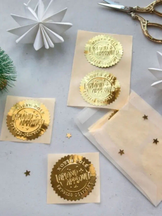 Embossed gold foil "North Pole Lapland Approved Express Delivery" seals on cream backing sheets. They sit beside a glassine envelope, gold confetti stars and a pair of gold scissors.
