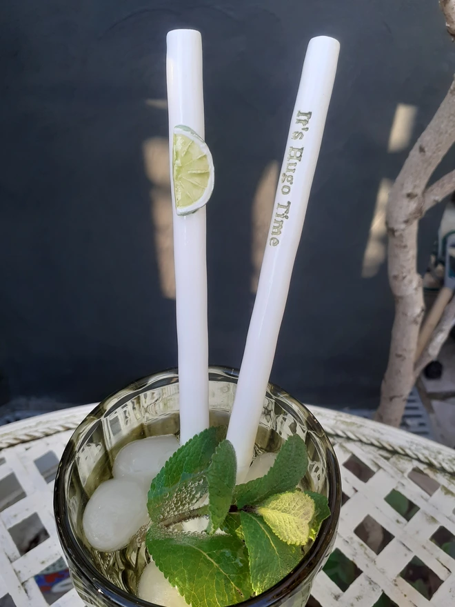 Handmade ceramic cocktail straws in a Hugo Spritz cocktail, one with a raised lime slice motif and one reading “It’s Hugo Time”, in a glass with ice and fresh mint.