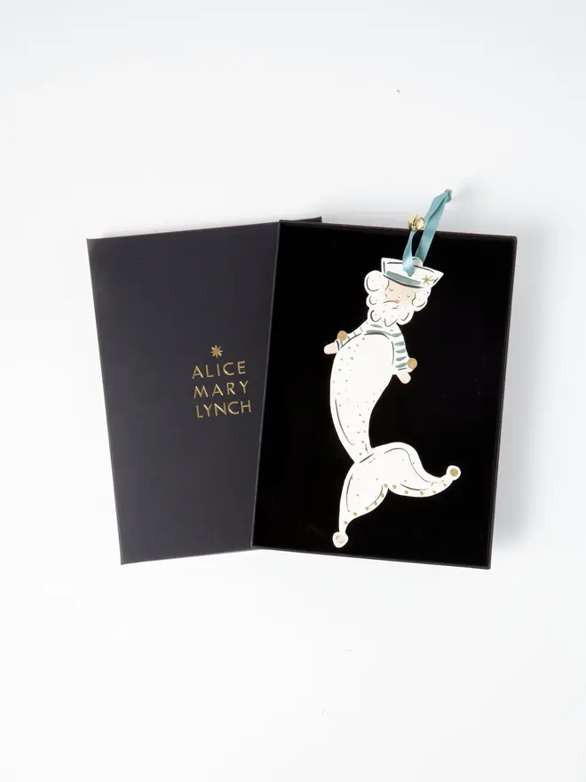 Merman Tree Dancer. the merman tree dancer hanging ornament sits inside its black presentation box, with the lid open to reveal "alice mary lynch" embossed in gold lettering. the bearded merman figure wears a sailor hat and striped teal top, with a cream scale-patterned tail edged in gold curling into a jester-style pointed tip. the branded box gives the piece a polished, gift-ready presentation.