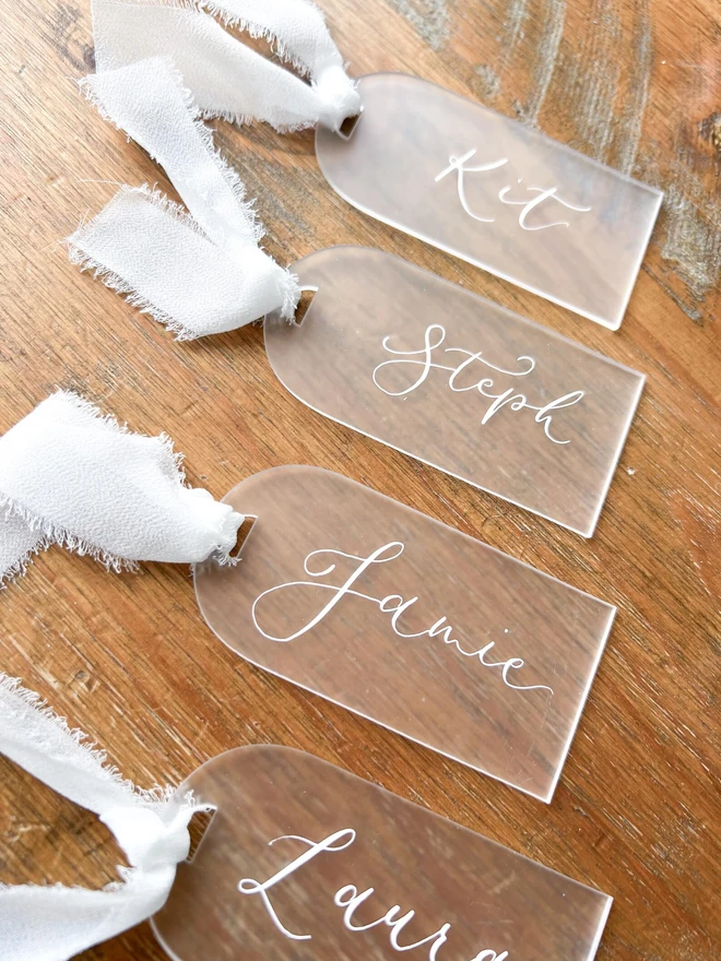 Personalised Calligraphy Frosted Acrylic Place Cards, frosted acrylic place cards with white chiffon ribbons grouped together on a wooden table. 