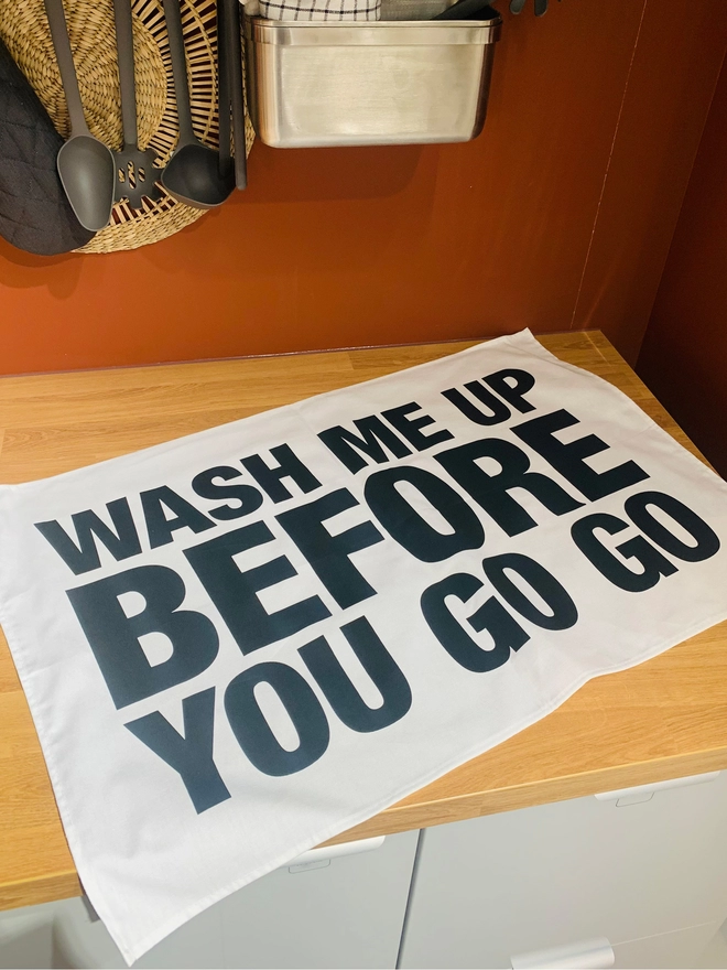 London Drying Wash Me Up Before You Go Go black text on white tea towel laying on kitchen counter with utensils hanging on back wall