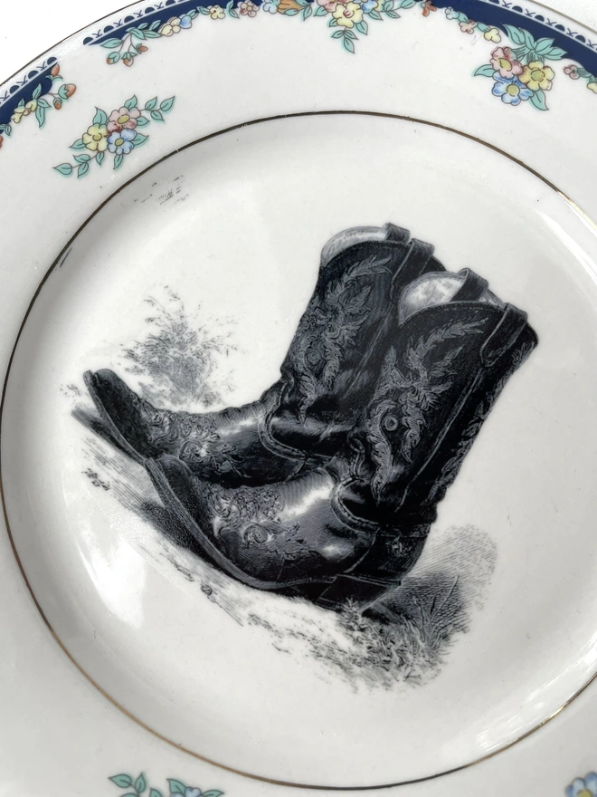 a vintage plate with an ornate border has a black and white vintage illustration of a pair of ornate cowboy boots in the centre