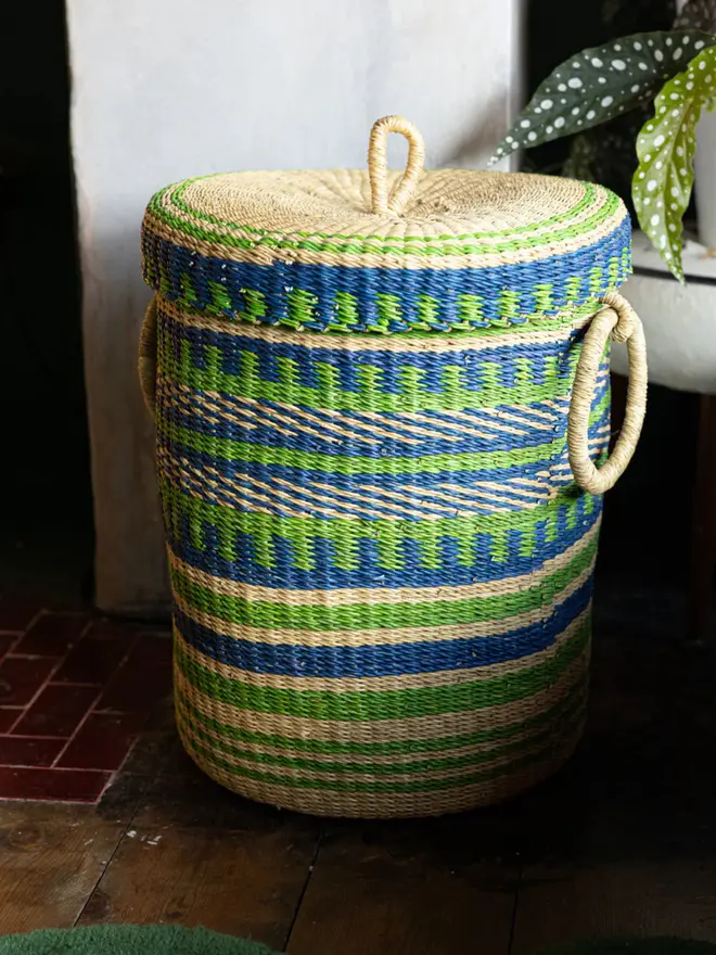 Ghanaian Hand Woven Laundry Basket With Lid And Handles, a colourful basket sitting on a wooden surface in a decorated living room. 