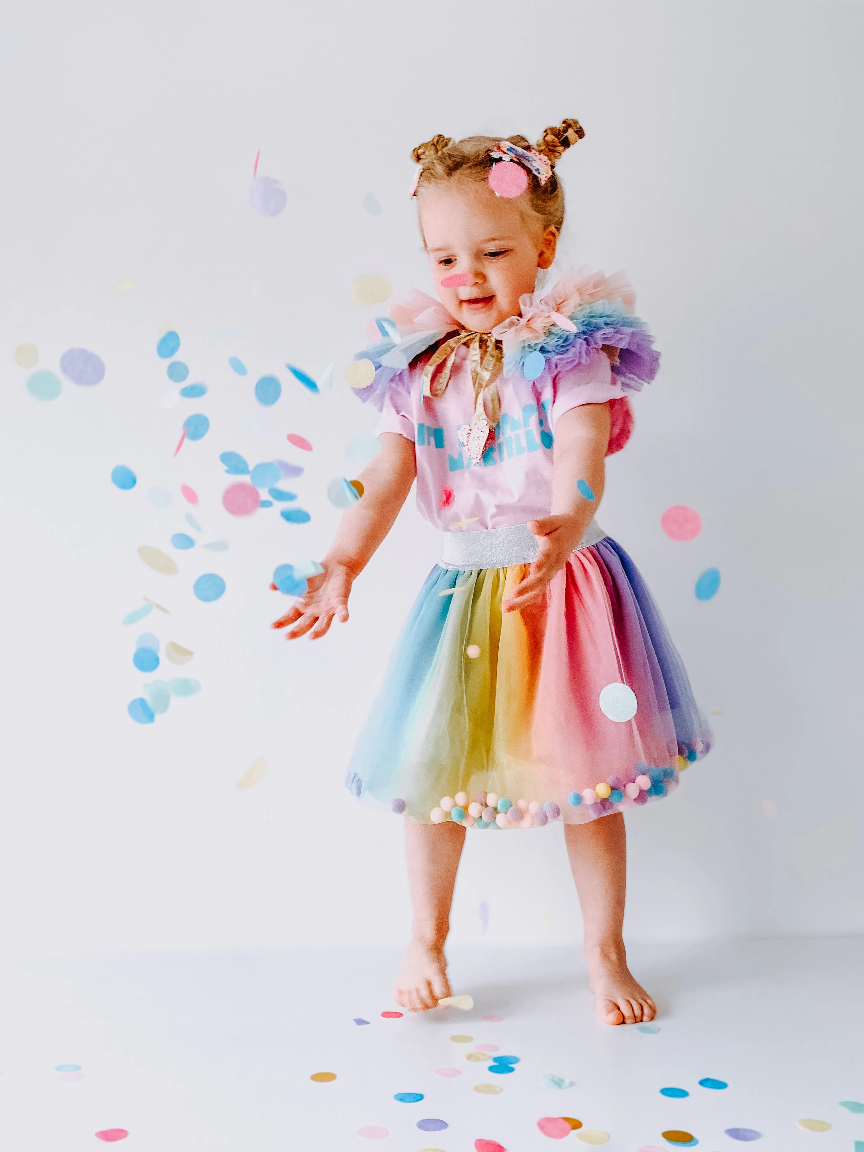 Child wearing a rainbow tutu filled with pom poms throwing confetti