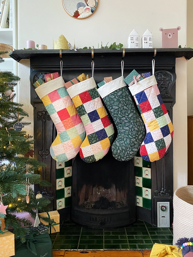 Four quilted patchwork Christmas stockings hang on a black fireplace, beside a large decorated Christmas tree.