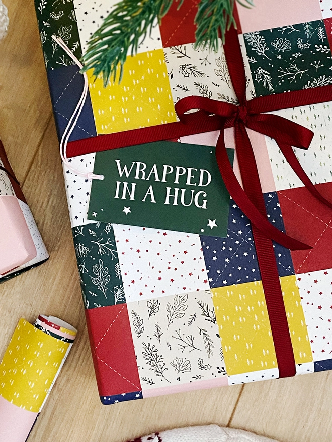 Gifts wrapped in a traditional patchwork design wrapping paper are on a wooden floor beneath a Christmas Tree.