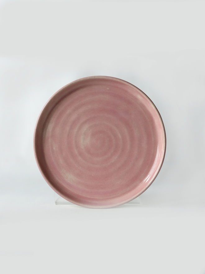 A handmade ceramic cake plate in blush pink glaze, photographed from the front against a plain light grey background, showing a natural concentric swirl texture and a shallow raised rim
