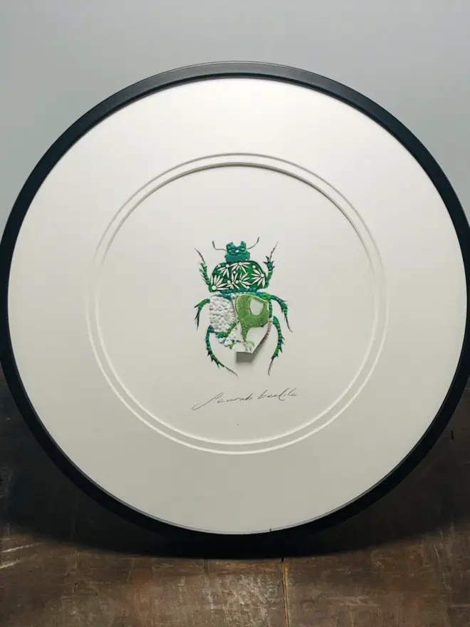 Hand-drawn, painted, mosaic clay, vintage green Dragon pottery insect on white double mounted card, in a black circular frame.
