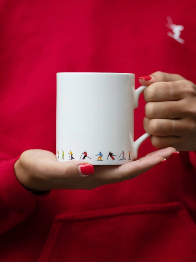 Ski Chain Mug. A simple white mug, the lower portion of which is illustrated with a pattern of multicoloured skiers. The mug is seen held against a red torso.