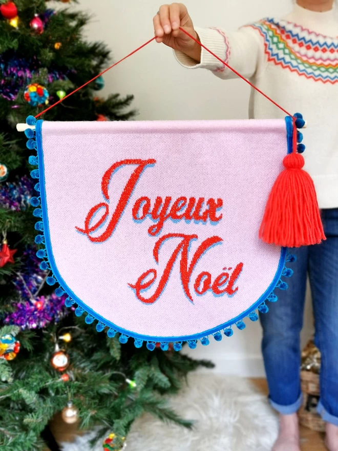 A woman wearing a colourful Christmas fairisle jumper stands next to a Christmas tree with retro coloured baubles. She is holding up a pink knitted scallop shaped banner with the words ‘Joyeux Noel’ written in a red retro text across it. The banner is trimmed with teal pom pom trim and has an oversize red tassel hanging from one side. 