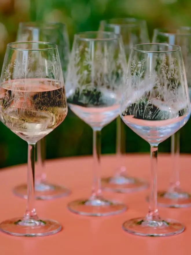 Meadow Wine Glass, a group of six etched wine glasses with wildflower patterns sit on a bright pink patio table. 
