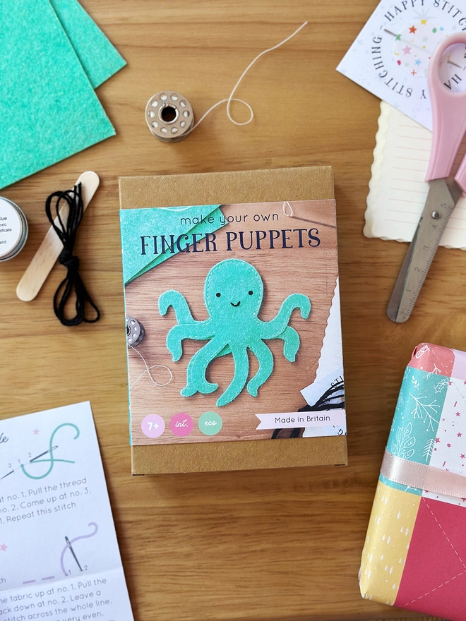 A small craft kit box to make a felt octopus finger puppet, lays on a wooden desk. Various craft kit contents lay around the box.