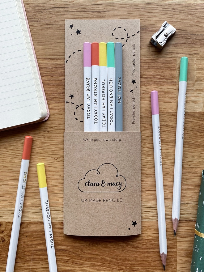 Five white pencils with pastel coloured ends and silver wording along the sides are tucked into cardboard packaging, on a wooden desk with four loose pencils and a notebook around them.