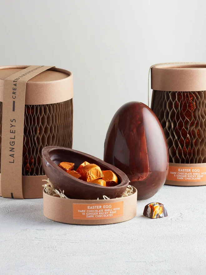 Large Dark Chocolate Easter Egg With Ginger Rocky Road, a chocolate egg sitting on a white surface. It is accompanied by another chocolate egg that is opened showing the chocolates within. 