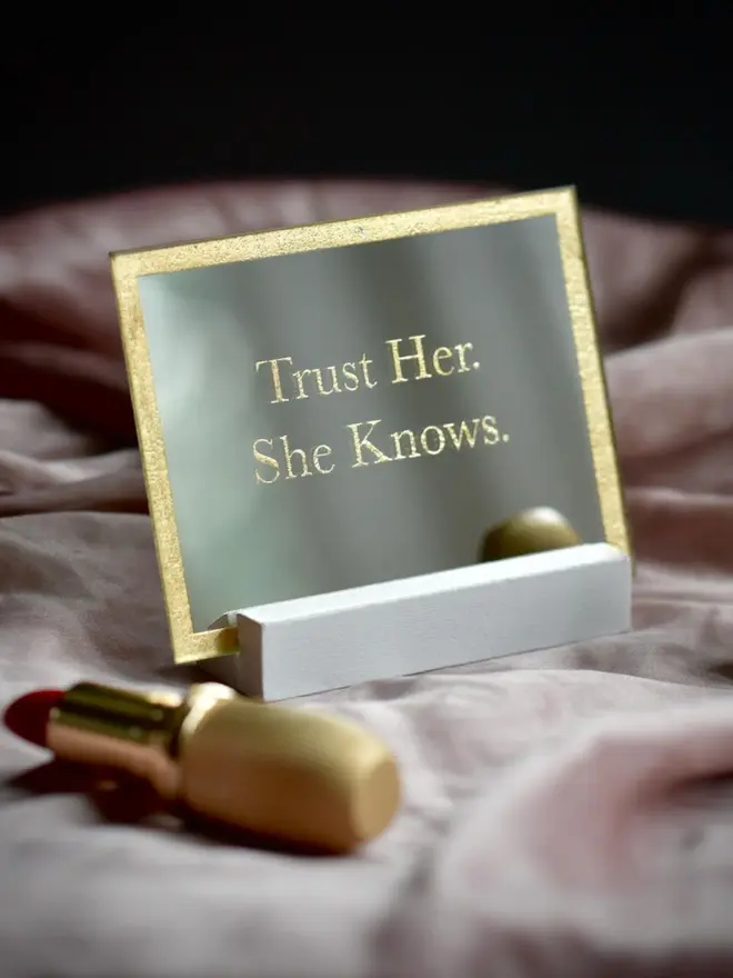 'Trust Her. She Knows' gold etched mirror on a white stand on pink silk