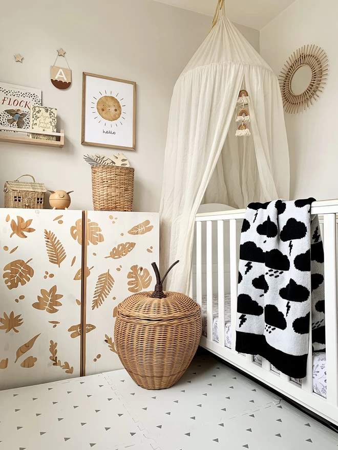 A neutral scandi style nursery with lots of natural wood, wicker accessories and a white cot. Over the cot is draped a black and white storm cloud blanket with a black trim.