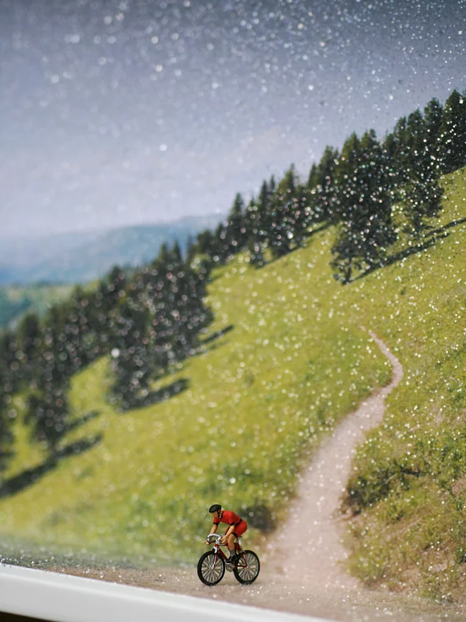 Miniature scene in an artbox showing a tiny man biking against a sparkling forested mountain backdrop.