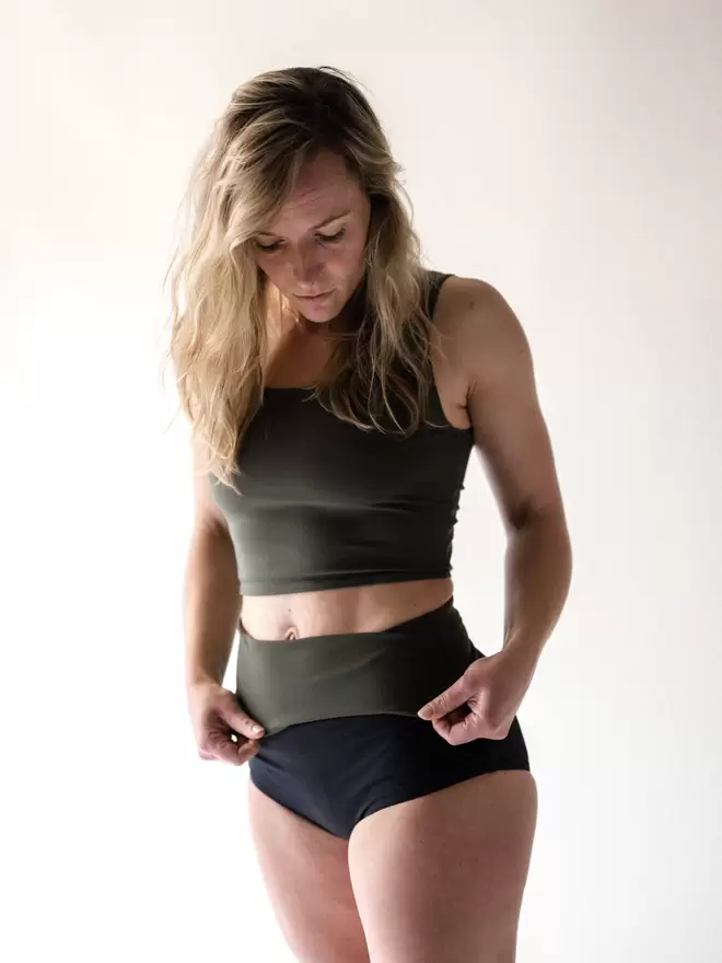 Blonde woman in studio looking down as she folds down her black high waist Davy J Sustainable Waterwear bikini briefs to show olive green lining, worn with olive green crop swim top