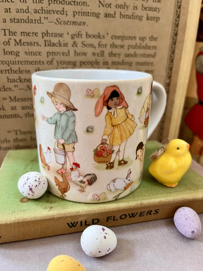 an illustrated easter mug the perfect size for a child or espresso for an adult featuring a traditional story book style illustration of sweet children doing easter activities easter egg hunt feeding chicks wearing an easter bonnet photographed next to mini eggs