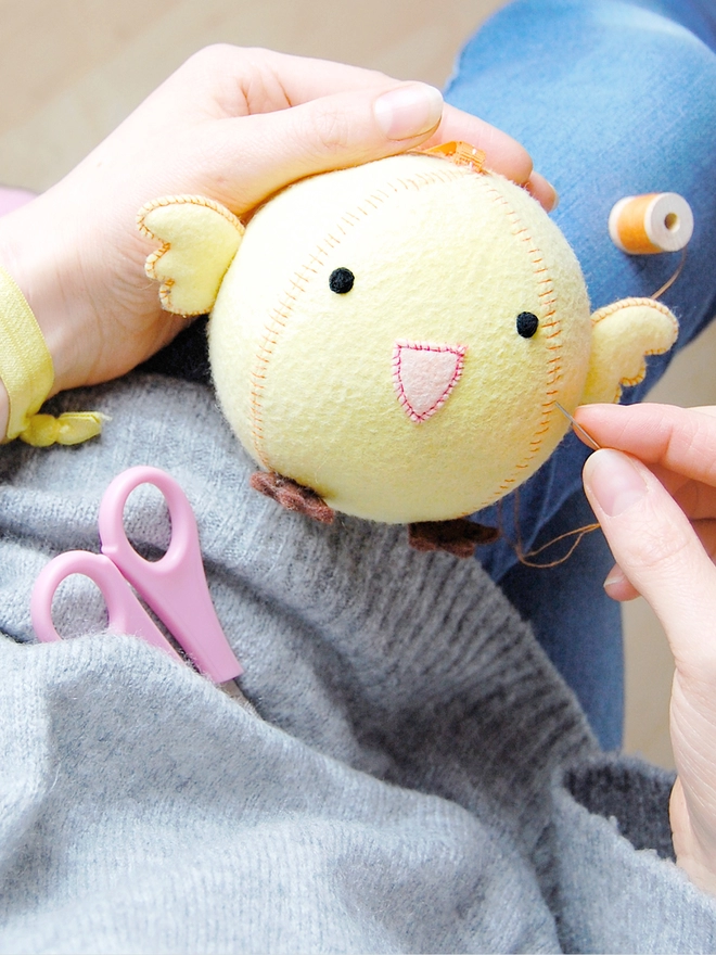 A round felt chick toy is being sewn together by someone wearing a grey jumper and blue jeans.