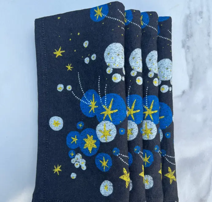 Solaris Napkins In White Or Blue. Set of four blue cloth napkins with blue and yellow starburst and orbital patterns, stacked in a staggered arrangement.