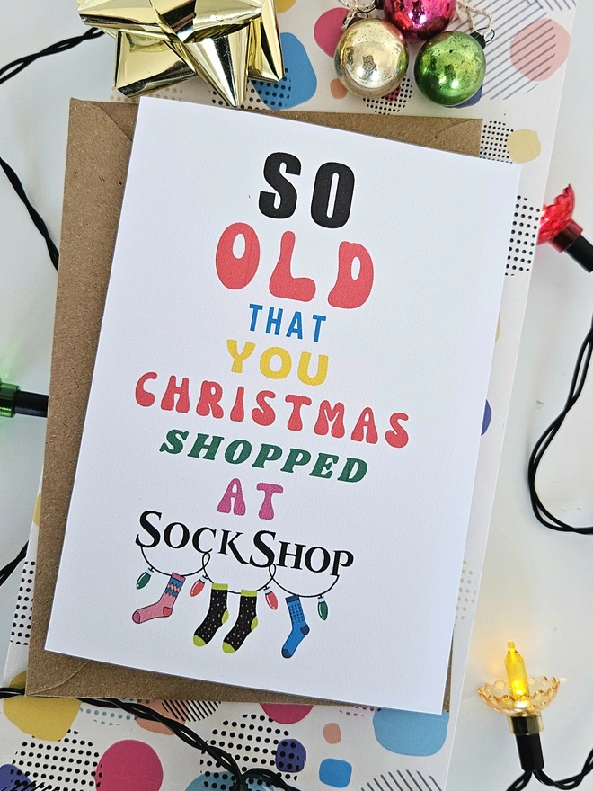 A photograph of the front of a humorous A6 greeting card illustrated using brightly coloured text and the retro shop logo. The text says so old you Christmas shopped at Sock Shop with 3 colourful socks hanging off a garland of lights