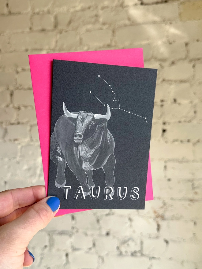 a greetings card featuring a greyscale illustration of a zodiac style bull with the text “TAURUS” with a neon pink envelope