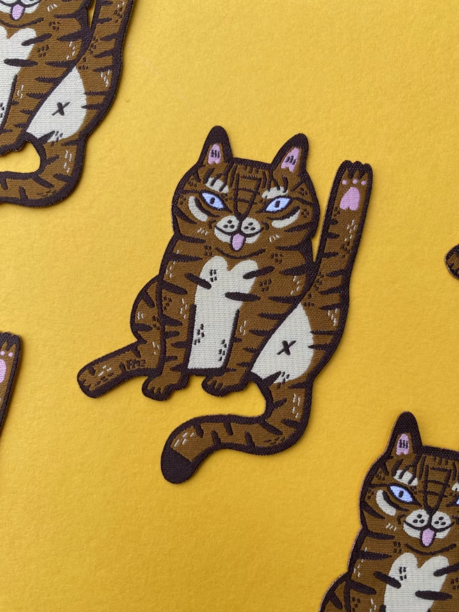Illustrated tabby cat licking themselves in the form of a woven iron-on patch 