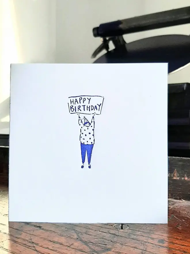 'Happy Birthday' Letterpress Handprinted Greetings Card, a white happy birthday card sitting on a wooden surface in a studio. It has a blue design. 