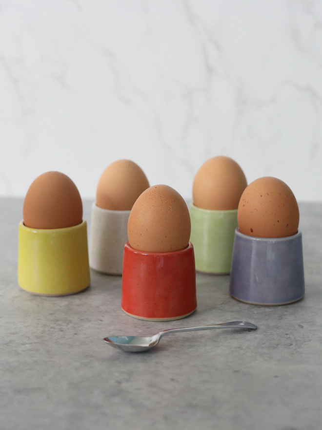 Five handmade ceramic egg cups each holding a brown egg, arranged on a grey marble surface in yellow, orange, purple, spring green, and cherry red glazes, with a small silver spoon in the foreground and a white marble background