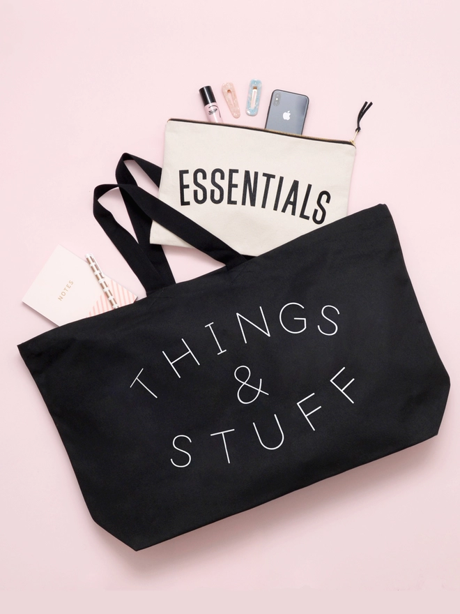 oversized black canvas tote bag with things and stuff slogan laying on a pink background with an essentials pouch spilling out