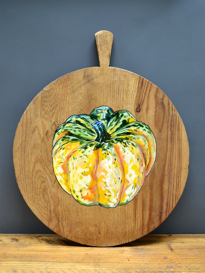 Wooden chopping board with handpainted design of a carnival squash standing against a wall