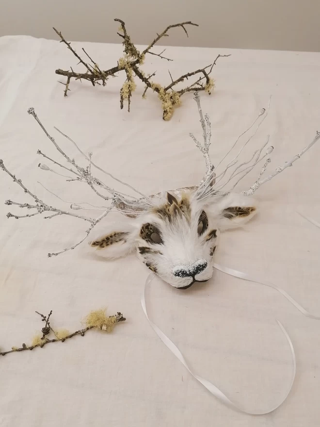 A white luxury deer part mask laying flat on a white cloth next to dried twigs