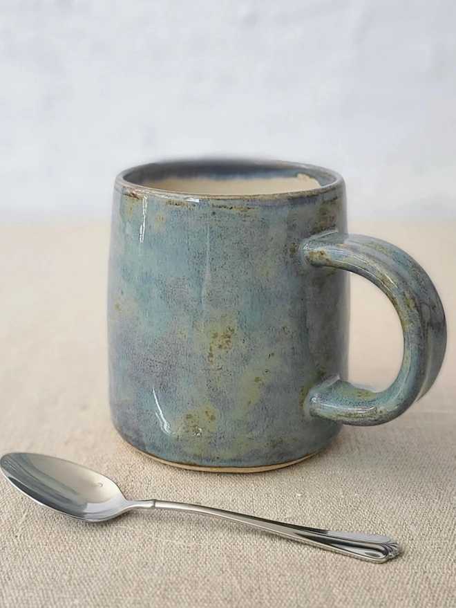 Ceramic Pint Mug, a colourful ceramic mug sitting on a plain surface against a white backdrop. It is accompanied by a spoon. 