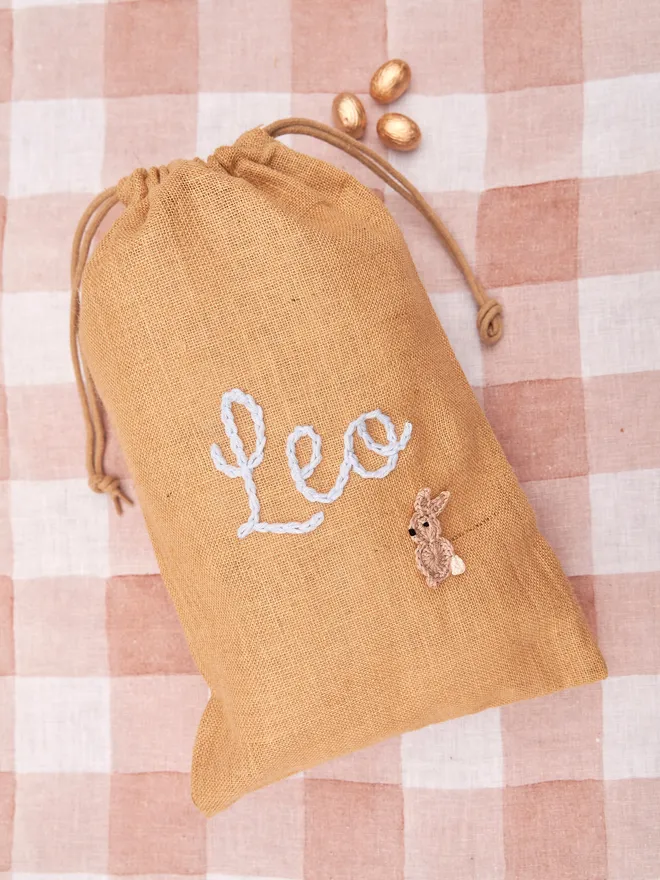 jute drawstring sack with brown knitted bunny decoration and blue 'leo' name next to golden chocolate eggs on red gingham table cloth