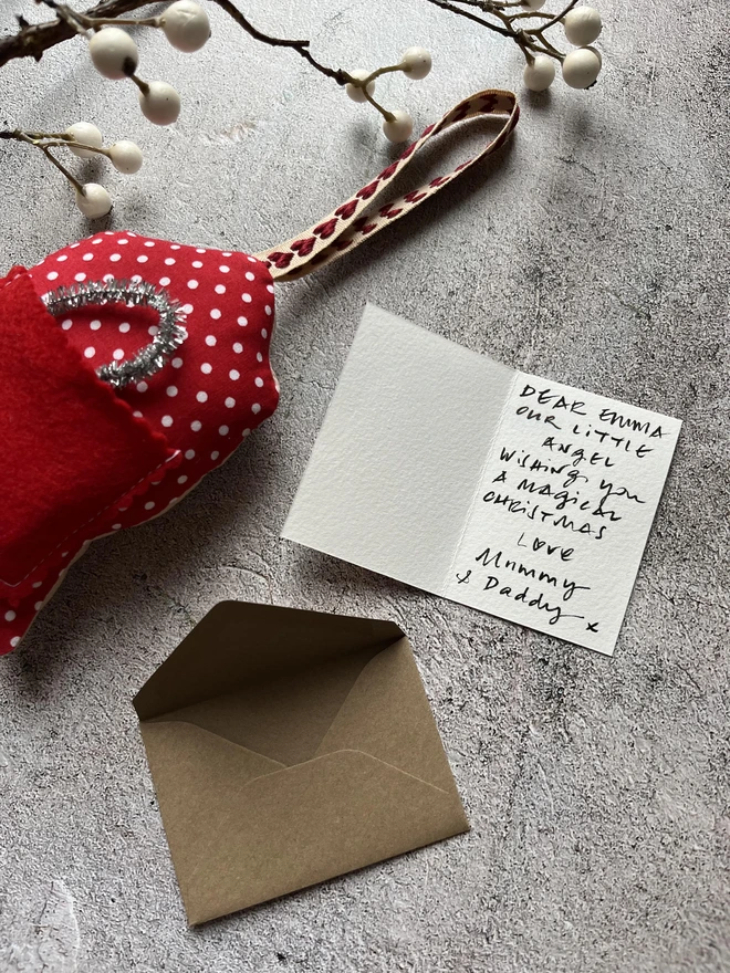 a photo showing a personalised minature greeting card wishing a little girl a magical christmas shown next to an angel christmas tree decoration with a red pocket on the back