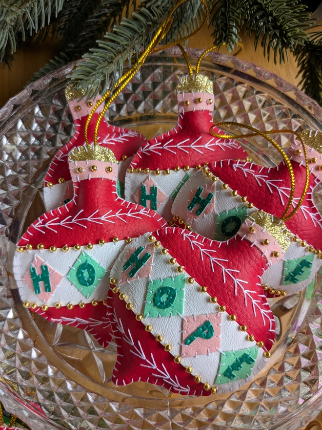 Collection of hand-stitched red, pink, green and white vegan leatherette Christmas bauble decorations spelling ‘HOPE’, embellished with gold beads, white embroidery and gold hanging loops, arranged in a vintage glass bowl.