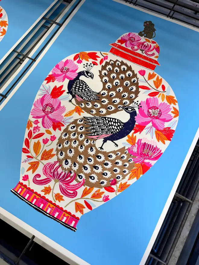 Peacock Vase Are You Looking At Me (You Probably Should), a colourful vase design on a blue screenprint, sitting against a studio rack. 