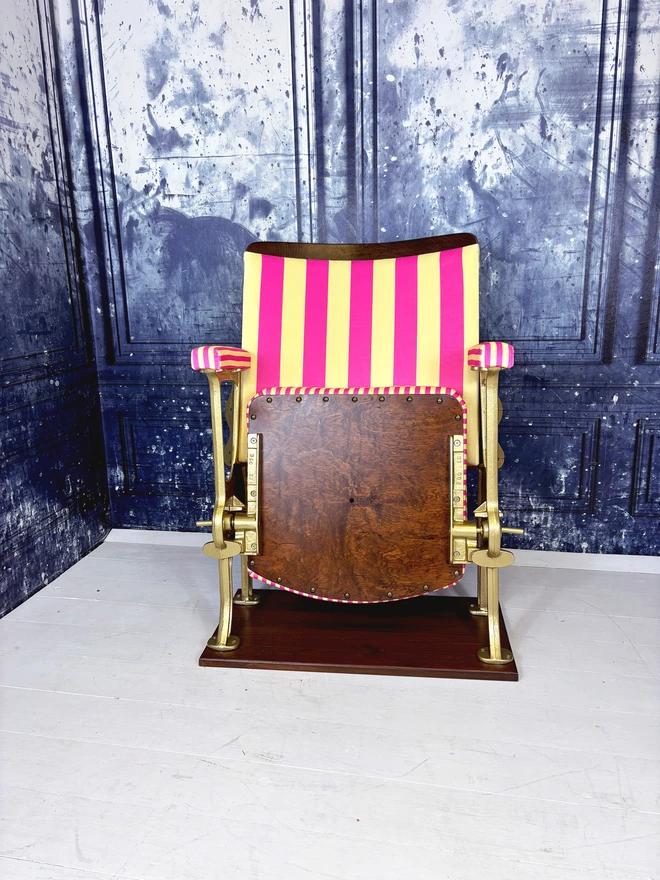 A pink and yellow vintage cinema seat.  The cinema seat has gold legs and is mounted onto a wooden plinth.  The fabric is pink and yellow striped.  The fold down part of the cinema seat is closed showing off the wooden seat base.