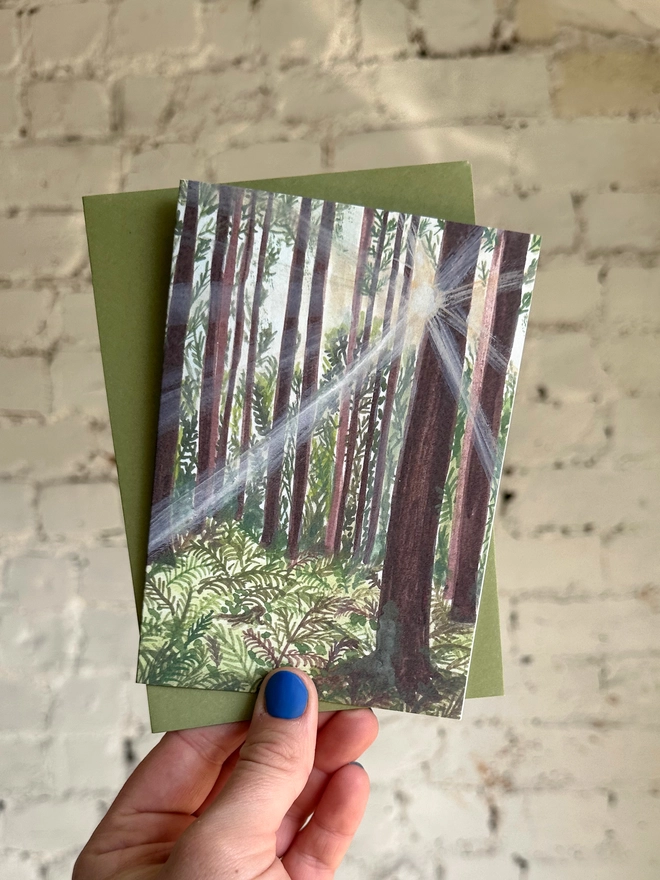 a greetings card featuring an illustration of sunlight beams coming through a woods with a green envelope