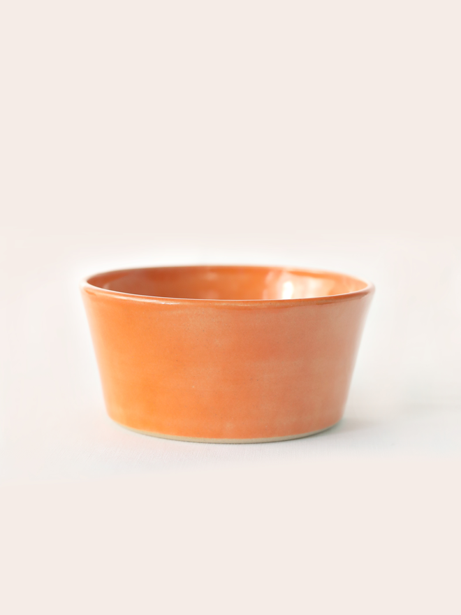 A single hand-thrown ceramic breakfast bowl in mandarin orange, with a glossy glaze showing subtle variation in tone, a gently flared form and unglazed natural clay base, photographed against a plain light grey background.