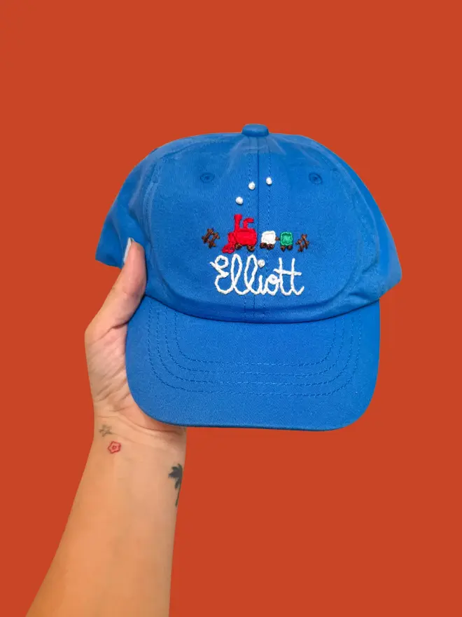 Hand Embroidered Kids Cap, a person holding a personalised cap against a colourful backdrop. It says a child's name. 