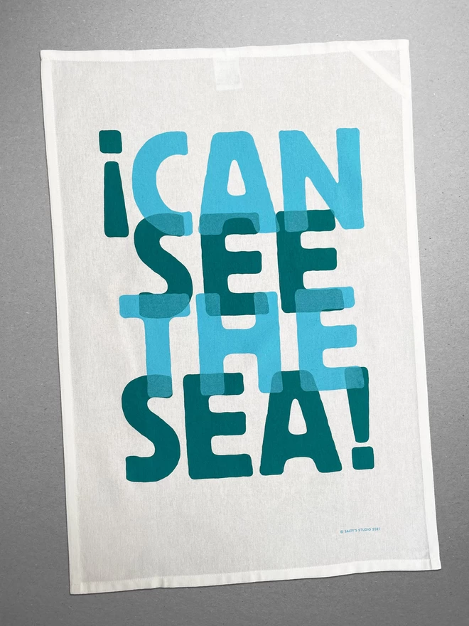 I Can See the Sea is screenprinted in two overlapping turquoise blues on this fairtrade cotton teatowel, spelling out the ubiquitous phrase, reminiscent of ocean waves. On a grey background. 